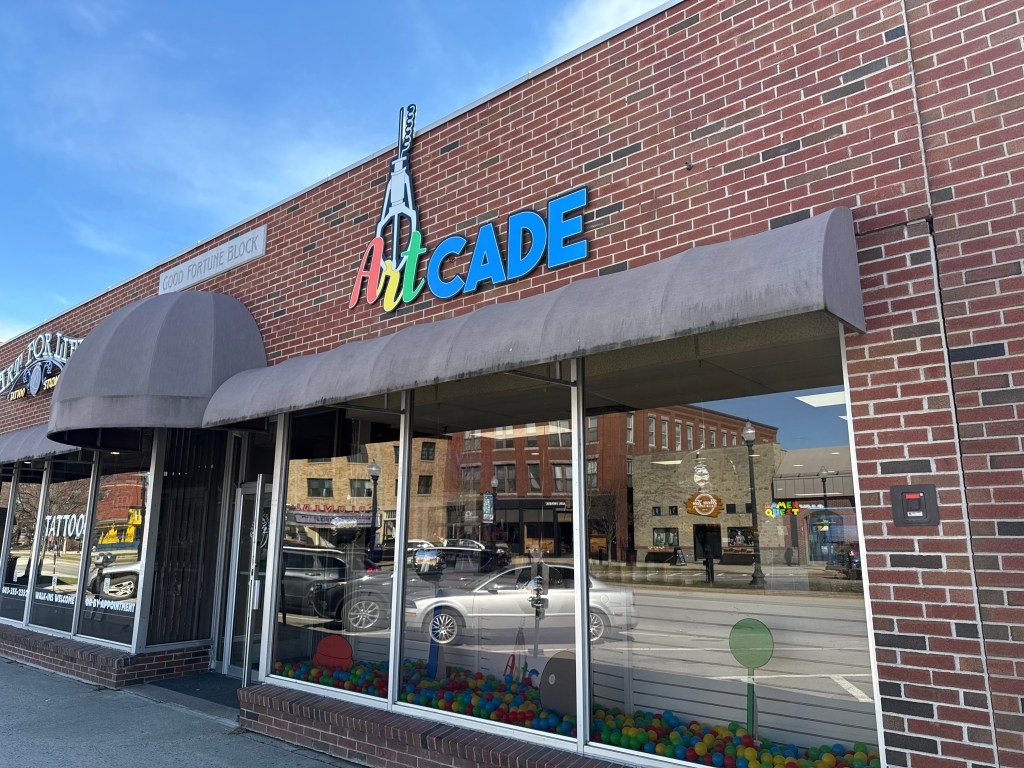 Exterior view of ArtCADE, a storefront with colorful signage against a brick building, featuring large windows displaying colorful balls inside.