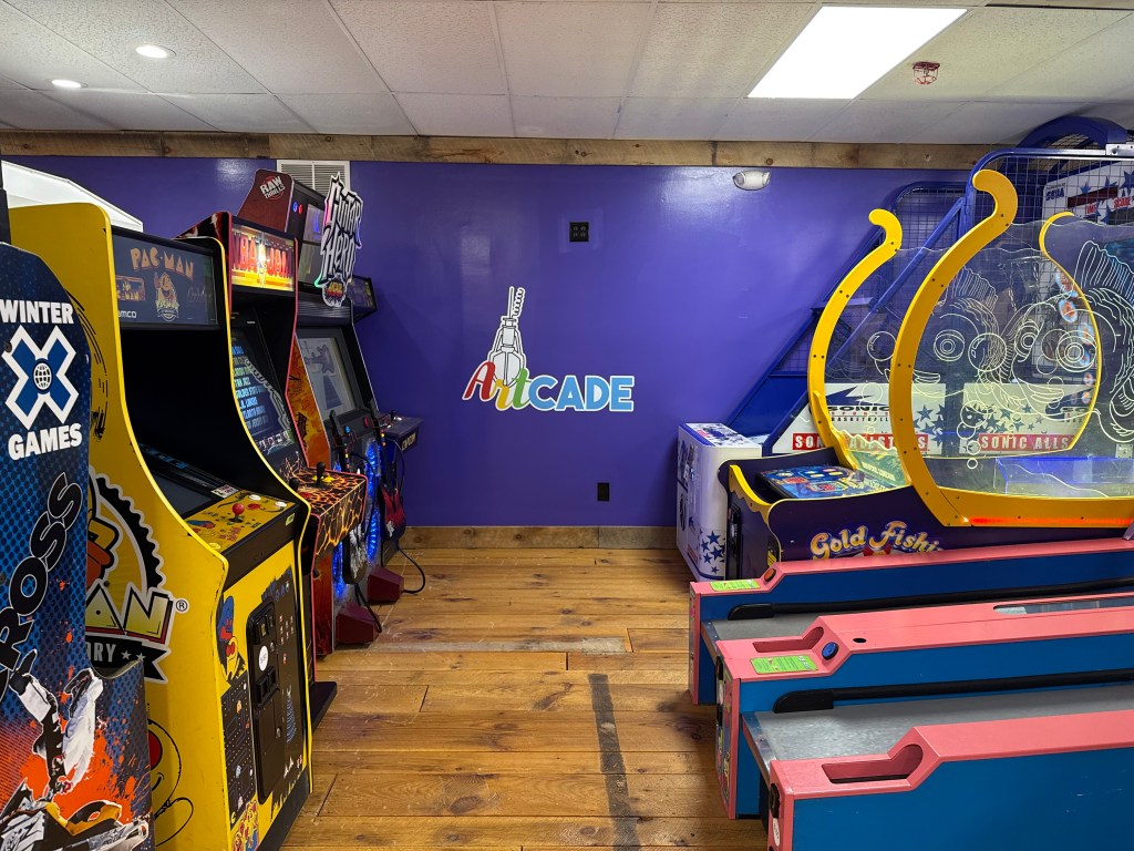 Interior view of an arcade featuring classic arcade machines, including Pac-Man, and various ticket redemption games. A colorful sign that reads 'Arcade' is displayed on the wall.