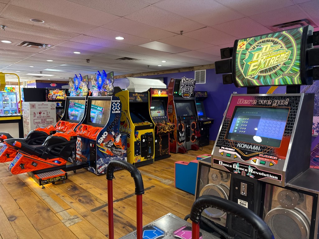 A vibrant arcade scene featuring various vintage gaming machines, including racing and dance games. Bright colors and illuminated screens create an inviting atmosphere.