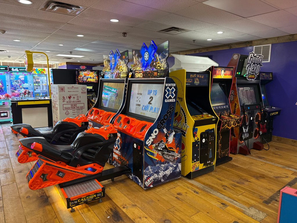 A row of arcade games in an entertainment center, featuring racing and classic game machines, with a wooden floor and colorful décor.