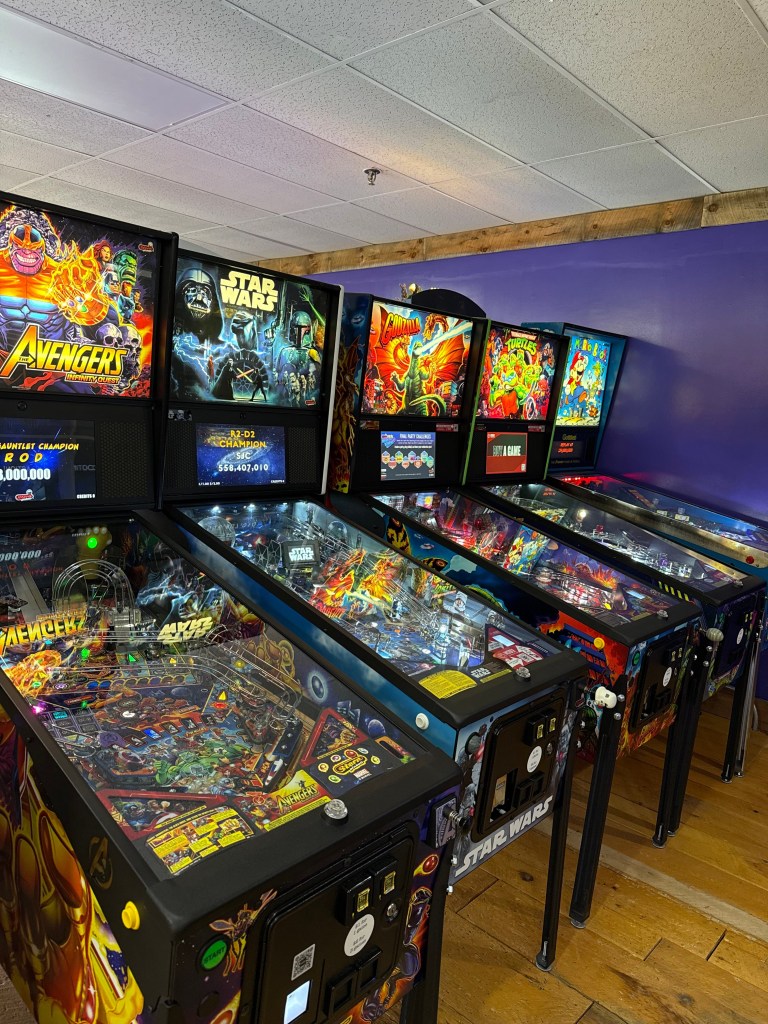 A row of colorful pinball machines featuring popular franchises like Avengers, Star Wars, and Godzilla, set against a purple wall in a gaming space.