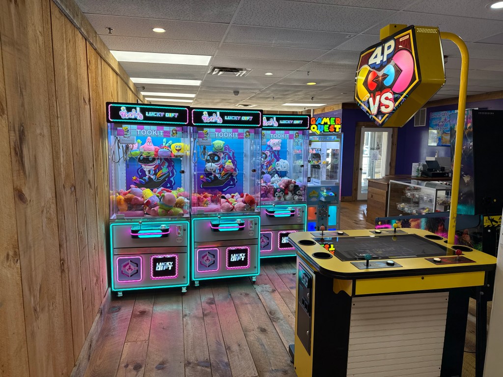 A vibrant arcade game area featuring two claw machines filled with colorful plush toys, alongside a multi-player arcade game station in a wooden interior.