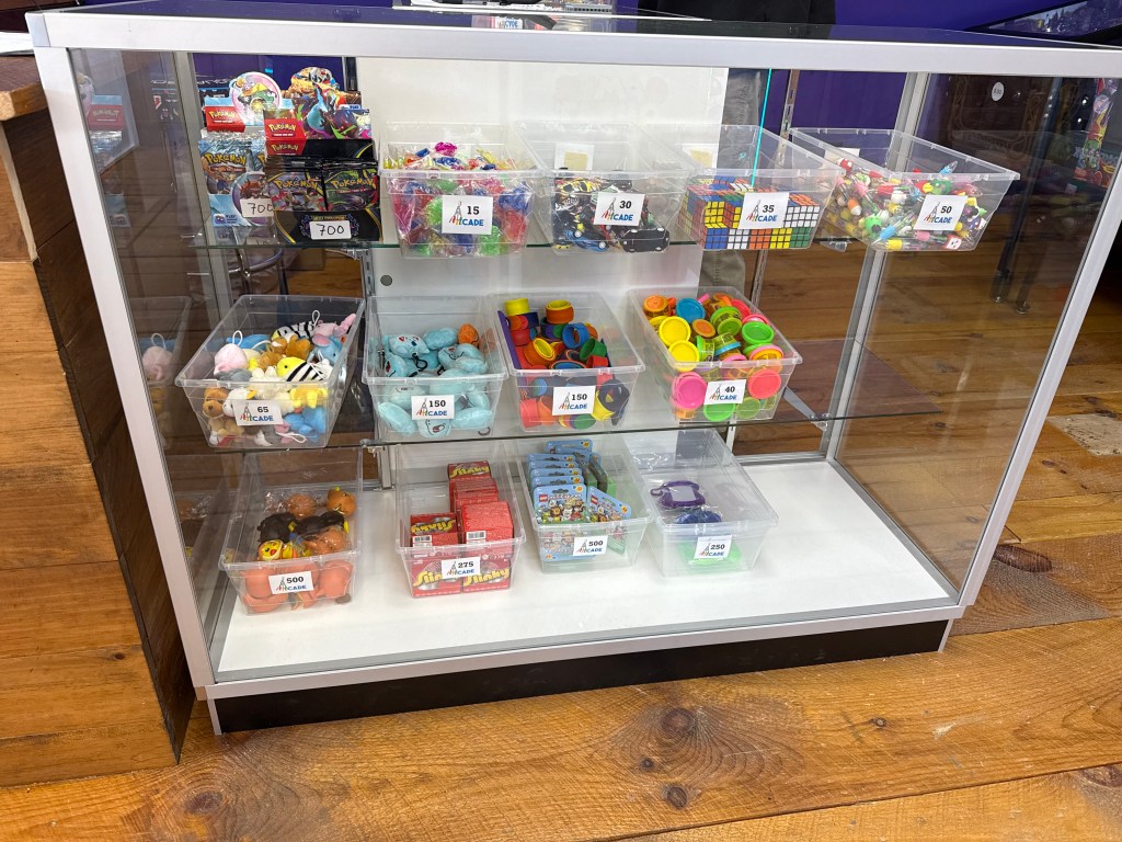 Display case featuring various toys and prizes organized in clear containers, labeled with prices, in a brightly colored arcade setting.