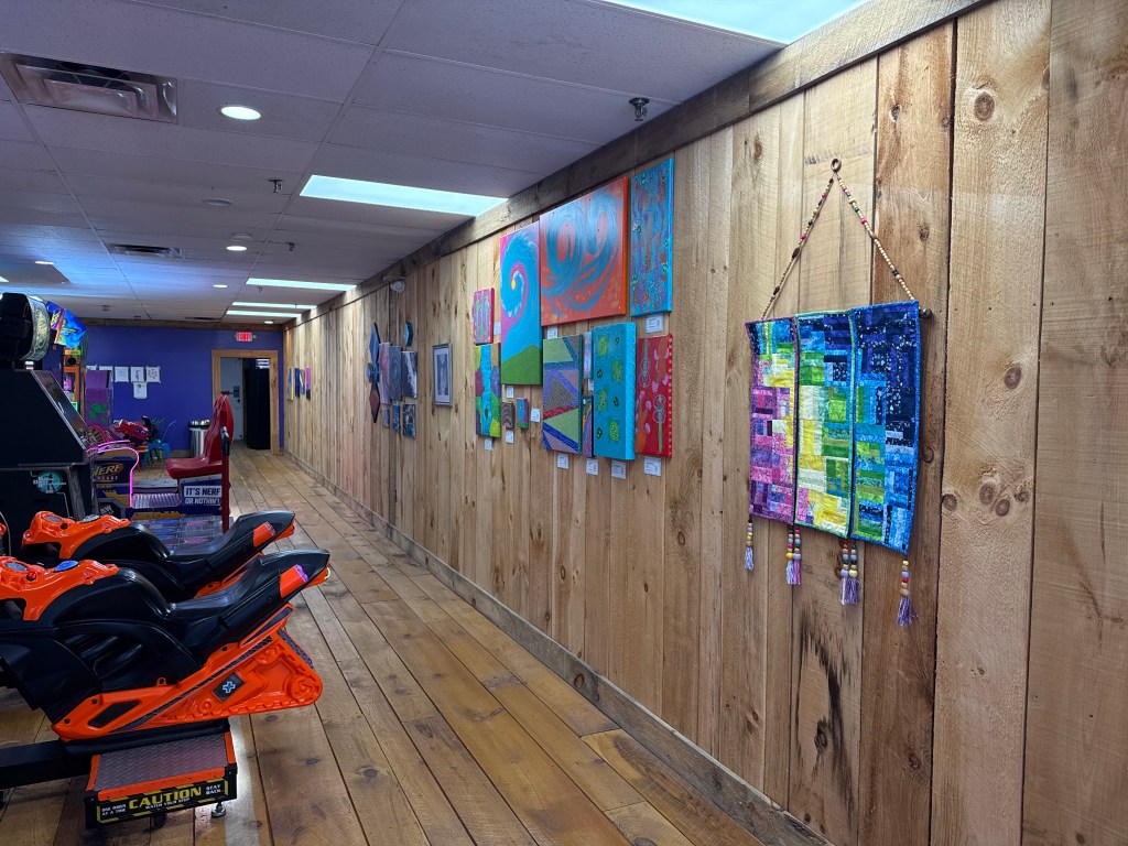 Interior view of a wooden-walled hallway featuring colorful artwork on display, with arcade game machines visible in the foreground.