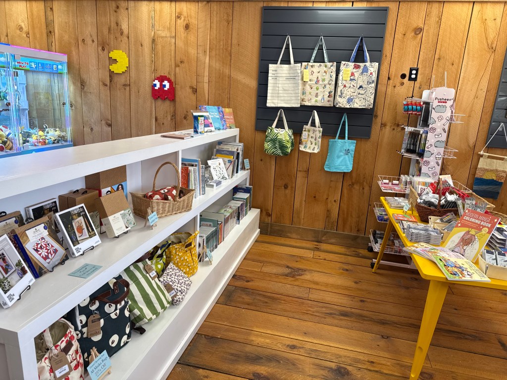 A cozy shop interior featuring wooden walls, displaying shelves with books and decorative items. On the left, colorful merchandise includes bags and stationery. A claw machine is visible in the background alongside pixel art decorations resembling classic video game characters.