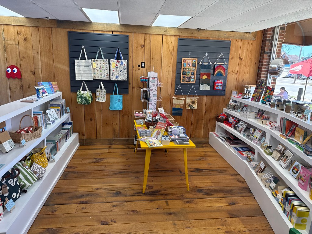 Interior view of a cozy retail shop featuring wooden walls, shelves displaying various books and items, and a bright yellow table in the center with products on it.