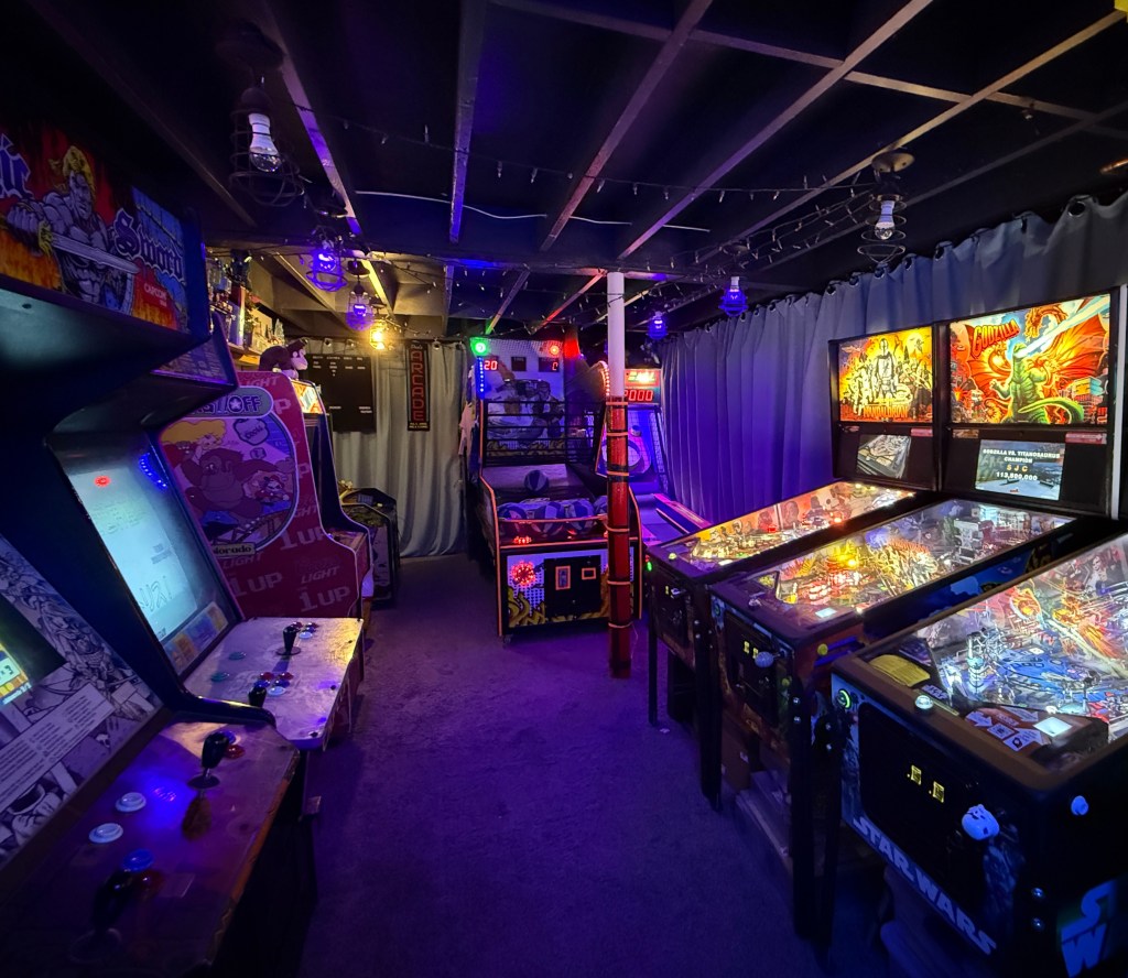 An indoor arcade with various vintage arcade games and pinball machines, illuminated by colorful lights.