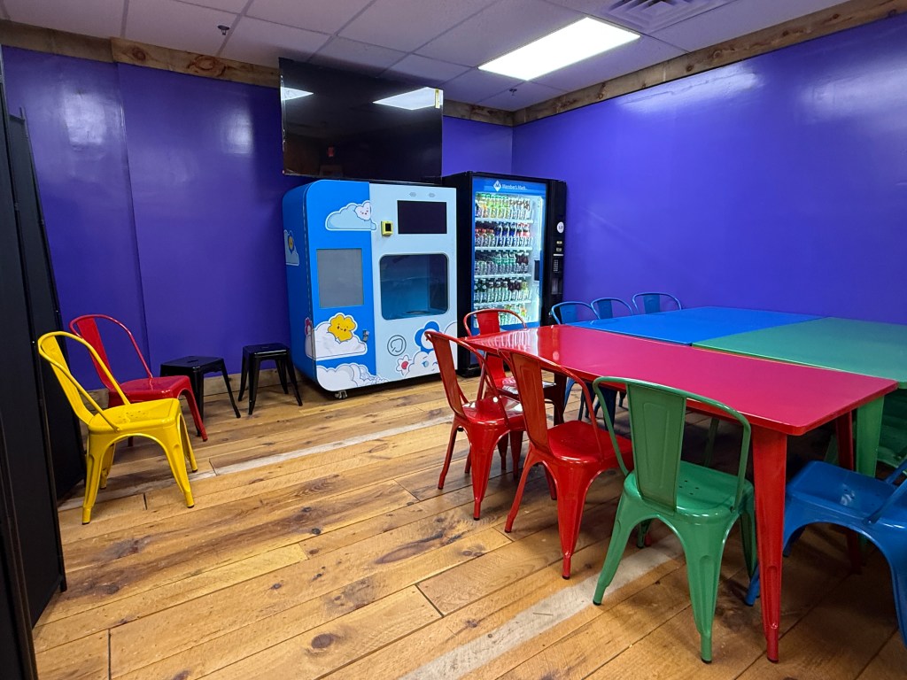 A colorful room with purple walls featuring red, green, yellow, and blue chairs surrounding vibrant tables. A vending machine and an arcade-style game machine are visible.