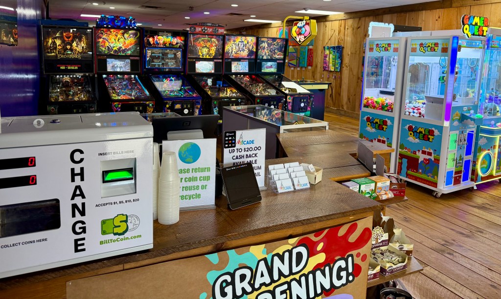 An indoor arcade featuring various pinball machines, a change machine, and claw machines, with a wooden interior and signage advertising a grand opening.