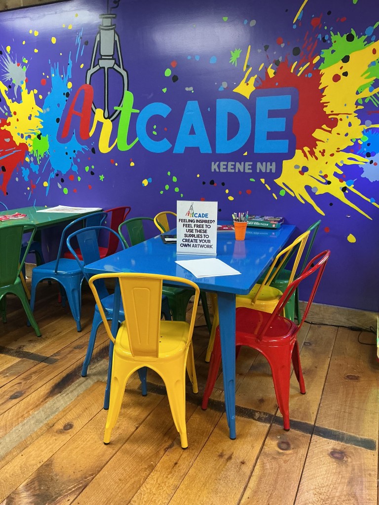 Colorful children's art space featuring a blue table surrounded by chairs in red, green, yellow, and blue, with a vibrant mural reading 'ArtCade Keene NH' in the background.